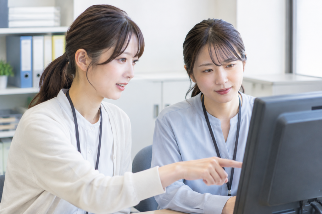 2人の女性がデスクでパソコン画面を見ながら指差しで確認している