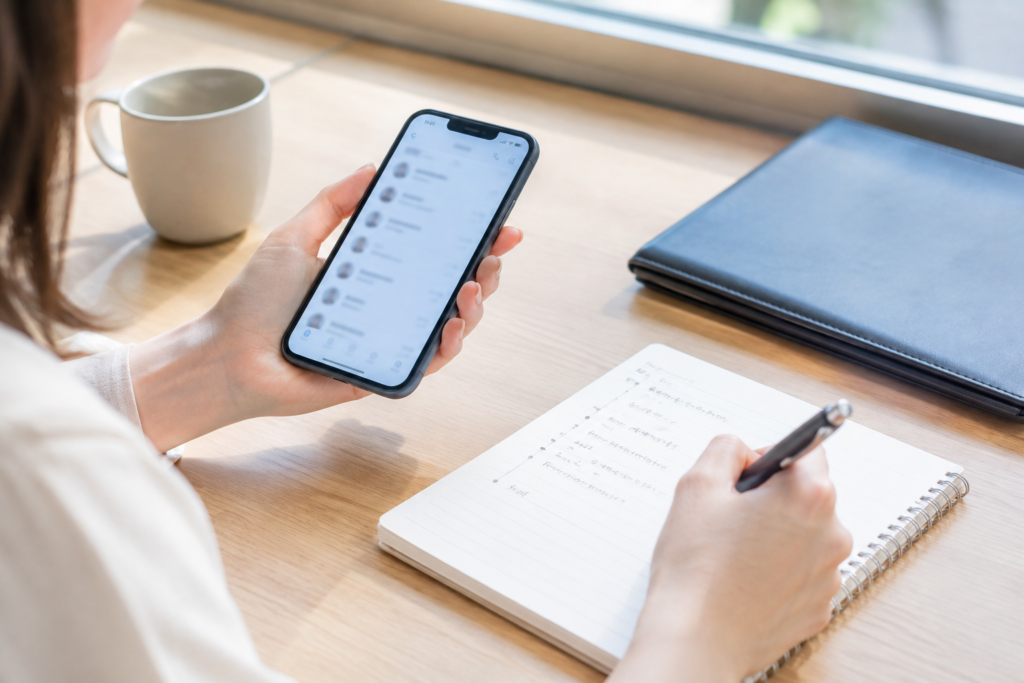 机の前でスマートフォンを見ながらノートにメモを書く人物